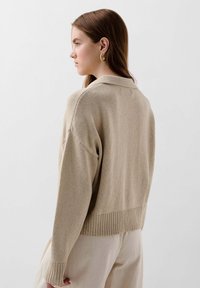 Beige gestrickter Pullover mit Schalkragen, gerippten Bündchen und einer verkürzten Saumlinie, der eine weiche Textur und einen lässigen Schnitt aufweist.