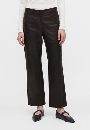 Personne portant un pantalon droit en cuir marron foncé, un haut blanc à manches longues et des ballerines noires pointues avec une bride et une boucle.