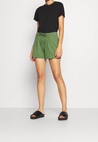 Zelené bavlněné kraťasy s elastickým pasem na šňůrku, spárované s černým tričkem s krátkým rukávem a černými klouzavými sandály. Hladký povrch.