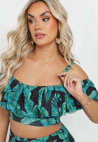 Svart off-shoulder crop top med volangdetaljer, prydd med ett livligt grönt lövmönster. Modellen bär blommiga örhängen.