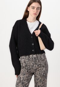 Cardigan nero lavorato a maglia con grandi bottoni, abbinato a pantaloni in stampa pitonata e una borsa beige. Il cardigan presenta una scollatura a V e una vestibilità ampia.