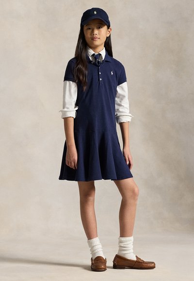 Ragazza in piedi che indossa un vestito blu navy sopra una camicia bianca con le maniche arrotolate, cappello blu navy, mocassini marroni e calzini bianchi arricciati su uno sfondo neutro.