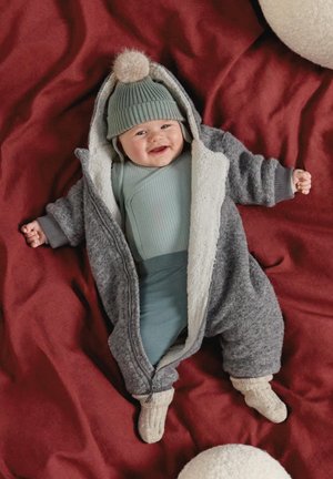 Bambino sorridente sdraiato su una coperta rossa che indossa una tuta grigia in pile, un completo verde a coste, un cappello abbinato con pon pon e calzini color crema.