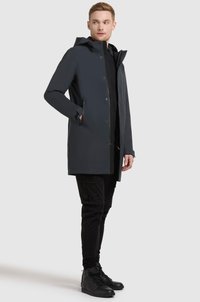 Donkergrijze waterdichte jas met capuchon, drukknoopsluiting en twee zijzakken, gecombineerd met zwarte broeken en zwarte hoge sneakers.
