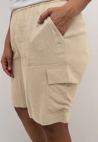 Beige cargo shorts dengan tali pinggang elastis, saku samping, dan kantong patch yang mencolok di paha. Terbuat dari kain berpola.