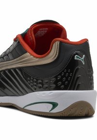 Chaussure synthétique noire avec une finition texturée, des accents dorés, un intérieur rouge et une semelle blanche ornée d'un motif vert et d'un logo intégré.