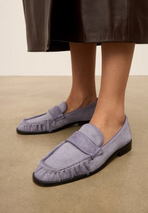 Pieds portant des mocassins pelucheux violet clair avec des semelles noires, debout sur un sol beige, associés à une jupe marron foncé.