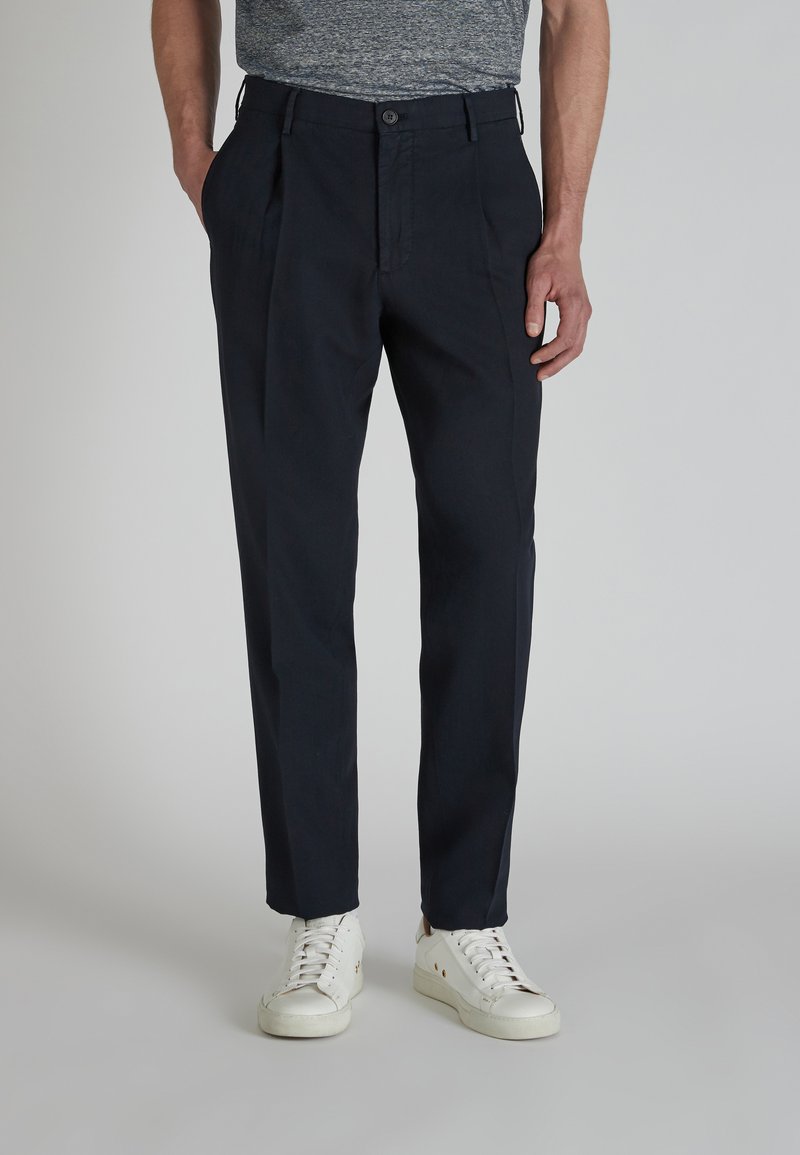 Pantaloni a pieghe blu navy in tessuto leggero con chiusura a bottone e design a gamba dritta, abbinati a sneakers bianche.