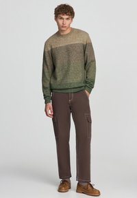 Maglione a righe marroni e verdi realizzato in materiale morbido, abbinato a pantaloni cargo marroni con molteplici tasche e cuciture a contrasto.