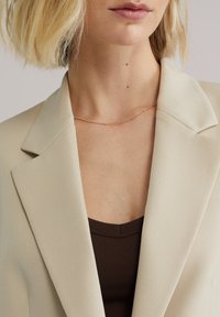Béžové sako so štruktúrovaným golierom, vrstvené cez tmavohnedý rebrovaný top. Zlatý jemný náhrdelník s malými okrúhlymi ozdobami.