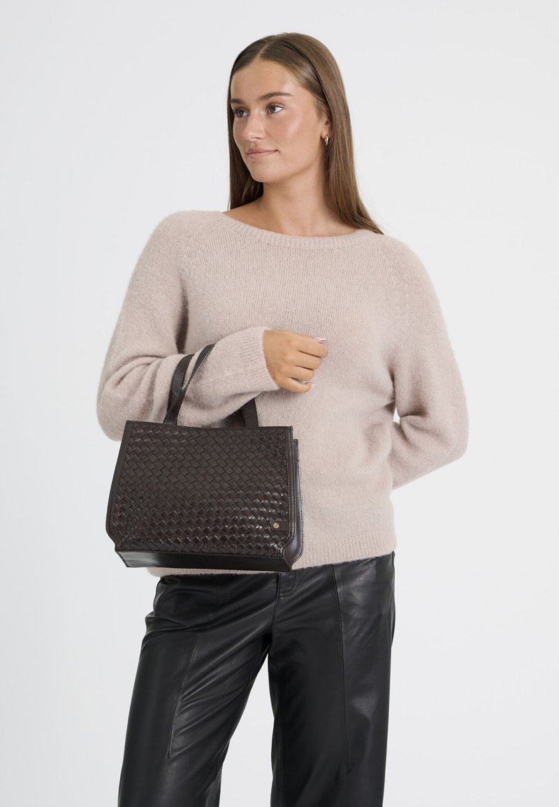 Jeune femme aux cheveux bruns, en pull beige et pantalon en cuir noir, tenant un sac à main tressé sombre avec deux poignées.