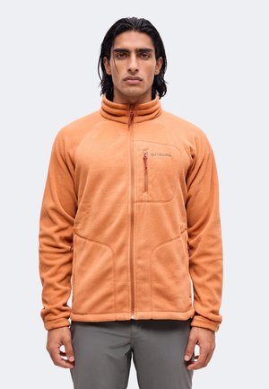 Mand med mørkt hår iført en orange fleecejakke med lynlås og grå bukser, stående med front mod kameraet foran en lys baggrund.