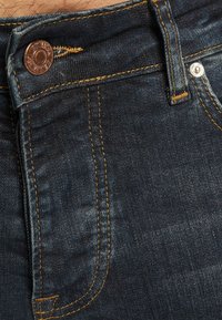 Tmavě modré džíny z denimu s texturovaným povrchem, kontrastní zlatým prošitím, zapínáním na knoflík a výraznou značkovou visačkou.