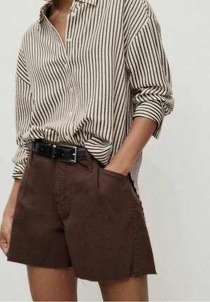Persona con camisa de rayas verticales en beige y negro metida en pantalones cortos marrones de talle alto con un cinturón negro.