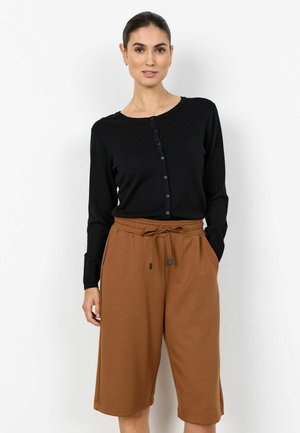 Naine seisab, kandes musta nööbitavat kardigani ja pruunid põlveni ulatuvad paelaga lühikesed püksid, taust on üksluine valge.