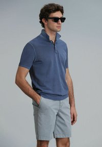 Blaues Baumwoll-Polohemd mit Kragen und kurzen Ärmeln, kombiniert mit hellgrauen Shorts. Das Modell trägt schwarze Sonnenbrille. Einfaches Design, lässiger Look.
