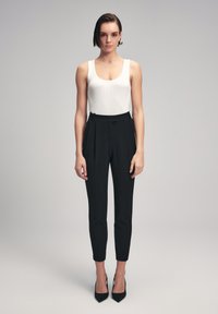 Witte geribbelde tanktop gecombineerd met zwarte hooggezaagde broek met plooien en taps toelopende benen, aangevuld met zwarte hoge hakken.