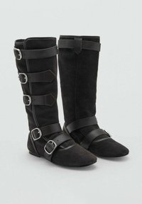 Botas de ante negras con una altura hasta la mitad de la pantorrilla, que presentan múltiples correas de cuero negro, hebillas plateadas y un diseño de punta afilada.