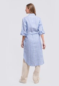 Striped blue and white linen shirt dress with rolled sleeves and a waist tie, featuring a longer back hem and lightweight texture.