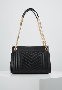 Borsa a tracolla in pelle nera con trapuntatura a zigzag, catene dorate e spallina in pelle nera, posizionata su una superficie bianca.