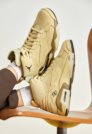 Beige høje sneakers med sort Jumpman-logo, båret med hvide sokker og brune bukser, hvilende på sædet af en træstol.