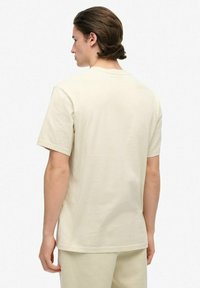 Hombre con cabello castaño de longitud media de pie de espaldas, vistiendo una camisa de manga corta beige clara y pantalones beige claros a juego.