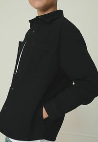 Schwarzes Button-Down-Hemd aus Baumwolle mit Kragen, Tasche auf der linken Brust und langen Ärmeln. Getragen über einem weißen Hemd und Jeans.