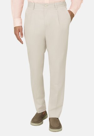 Beżowe, dopasowane spodnie noszone z brązowymi wsuwanymi butami i jasnoróżową, długorękawową koszulą, ręce spokojnie opuszczone wzdłuż ciała.