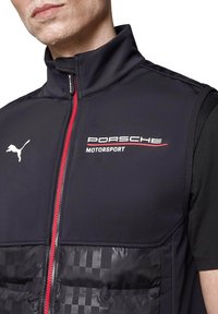 Czarna bezrękawnikowa kurtka z wysokim kołnierzem. Ma czerwony zamek błyskawiczny, logo "Porsche Motorsport" oraz teksturowaną, pikowaną dolną część z wzorem w kratę.