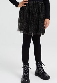 Svart glittrig tylkjol med veck, bärs över svarta ogenomskinliga strumpbyxor och i kombination med svarta snörkängor med mönstrade snören.