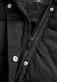 Veste légère noire avec fermeture éclair à l'avant, boutons pression et texture froissée. La fermeture éclair est noire avec des accents métalliques.