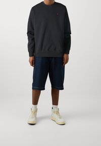 Sudadera negra de cuello redondo con un pequeño logotipo rojo, combinada con pantalones cortos de mezclilla y zapatillas altas blancas con acentos claros.