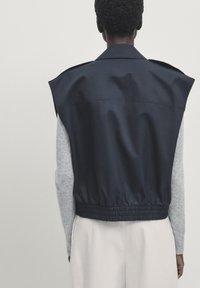 Granatowa, bezrękawnikowa kurtka z wysokim kołnierzem, elastycznym dołem i gładką fakturą, noszona na jasnoszarej bluzce z długim rękawem.