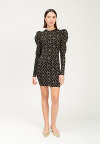 Abito nero con motivo geometrico dorato, maniche lunghe aderenti e spalle a sbuffo. Il tessuto ha una texture liscia. Abbinato a ballerine a punta.