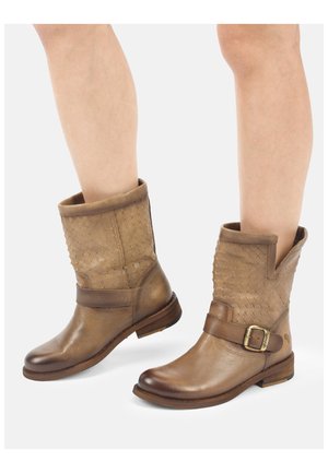 Benen met beige lederen laarzen tot halverwege de kuit, met een gestructureerd bovenwerk, band en metalen gesp, afgebeeld tegen een effen witte achtergrond.