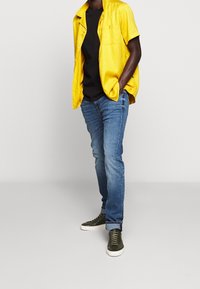Camicetta gialla a maniche corte con bottoni sopra una t-shirt nera, abbinata a jeans azzurri chiari e sneakers verde scuro. Sfondo semplice.