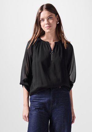 Schwarze, halbtransparente Bluse mit Rundhalsausschnitt und kurzen, ballonähnlichen Ärmeln. Verfügt über einen Reißverschluss und eine weiche, fließende Textur. Getragen mit dunklen Jeans.