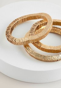 Set med tre armband i guldfärg med en strukturerad, rektangulär design. Varje armband har en polerad yta och varierande storlekar, staplade på en vit cirkulär bas.
