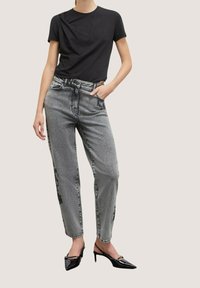 Černé tričko s krátkým rukávem a texturovanými detaily, spárované s šedými džíny s vysokým pasem zdobenými květinovým vzorem a černými špičatými podpatky.