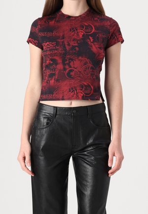 Kobieta w czerwono-czarnym wzorzystym krótkim T-shircie i czarnych skórzanych spodniach z wysokim stanem na jednolitym tle.