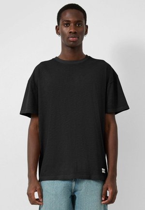 Młody mężczyzna z krótkimi włosami i kolczykami, ubrany w prostą czarną, luźną koszulkę i jasnoniebieskie dżinsy, stojący na białym tle.