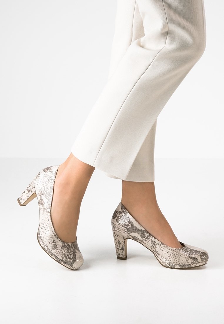 Femme portant des chaussures à talons épais à motif peau de serpent beige et gris avec un pantalon blanc raccourci sur fond blanc.