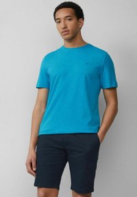 Türkises T-Shirt mit kurzen Ärmeln in glatter Textur; zeigt ein dezentes Logo auf der Brust. Kombiniert mit dunkelblauen Shorts, was einen lässigen Look kreiert.