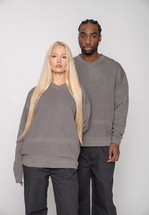 Deux adultes debout côte à côte portant des pulls gris côtelés assortis et un pantalon noir, devant un fond uni.