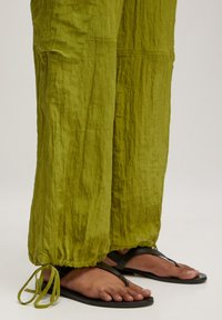 Groene, gestructureerde, loszittende broek met verstelbare trekkoorden bij de enkels, gecombineerd met zwarte flip-flops.