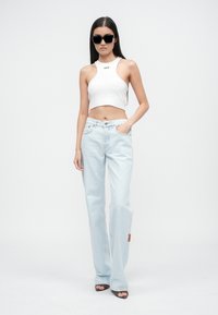Top corto blanco con el texto "OFF", jeans de pierna ancha azul claro y gafas de sol negras; modelo de pie en un fondo neutro.