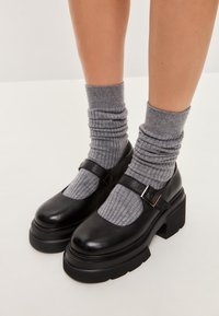 Pantofi Mary Jane negri din piele cu o talpă groasă, având o curea și o cataramă. Purtați cu șosete gri cu textură, care prezintă un tricot texturat.