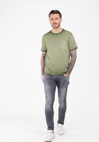 Hombre con brazos tatuados que lleva una camiseta estampada color verde oliva, jeans ajustados grises desgastados y zapatillas blancas, de pie con una mano en el bolsillo.