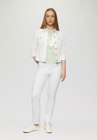 Weiße Denimjacke über einem grünen T-Shirt mit großem weißem Logo, kombiniert mit weißen Jeans und hellen Sneakers, die ein einfaches Design aufweisen.