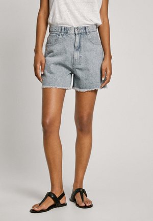 Kvinde iført lyseblå, frynsete denimshorts, hvid ærmeløs top og sorte flade sandaler, stående foran en neutral baggrund.